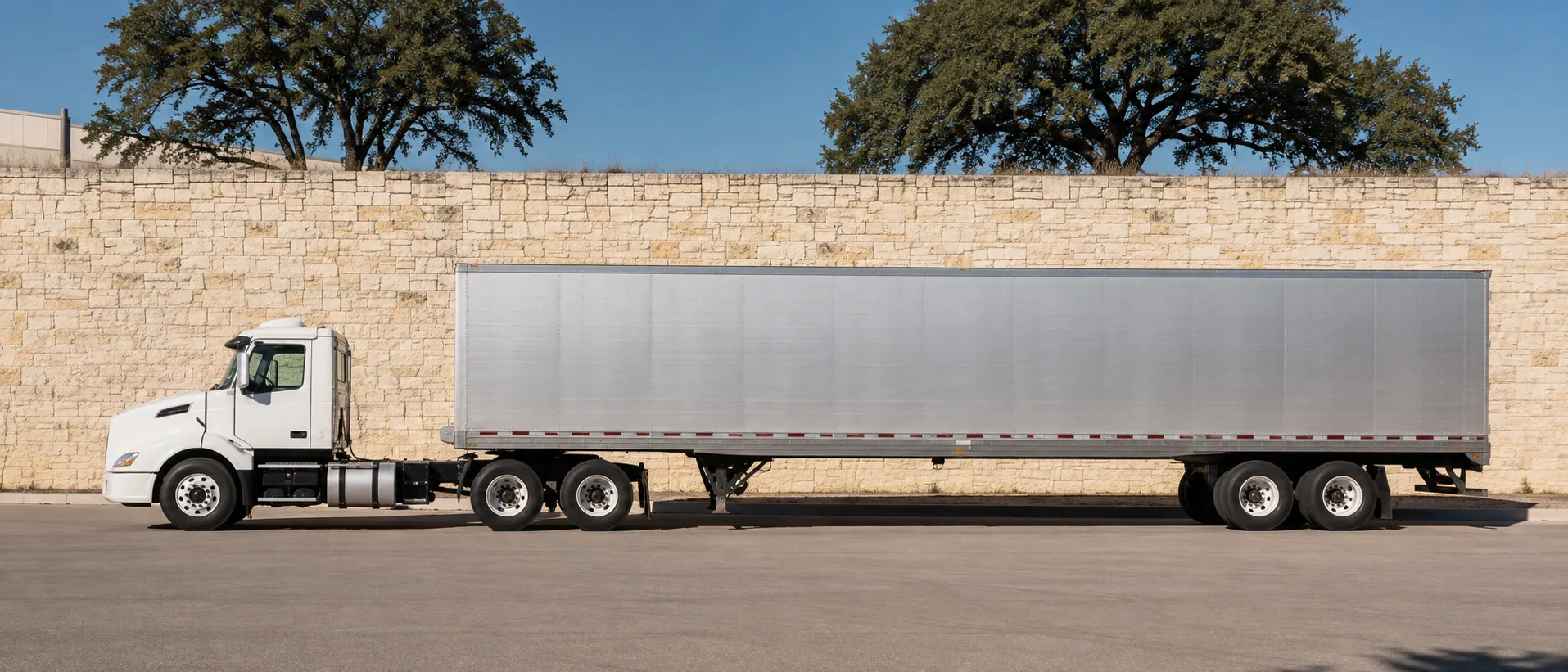 Illustrative side view of an unbranded tractor-trailer beside a pale limestone wall and live oaks.